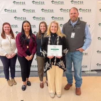 Group of five adults, four female and one male, standing in front of a CMCSS backdrop. Female with blond hair wearing a black sweater and cream colored pants is holding a certificate.